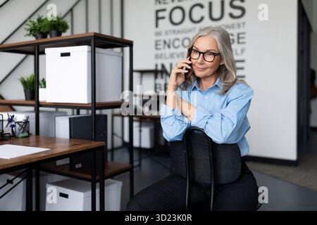 Grauhaarige ältere Geschäftsfrau mittleren Alters, die im Büro telefoniert, arbeitet mit Einwänden Stockfoto