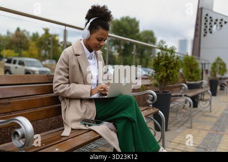 Afroamerikanische junge Studentin, die online an einem Laptop mit Kopfhörern arbeitet und auf einer Bank in der Nähe des Geschäftszentrums sitzt Stockfoto