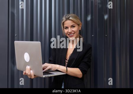 Porträt einer lächelnden Geschäftsfrau mit einem Laptop in den Händen vor dem Hintergrund einer grauen Bürowand Stockfoto