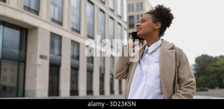 Junge erfolgreiche Versicherungsagentin mit afroamerikanischem Aussehen spricht auf einem Handy vor dem Hintergrund eines Anbieters Stockfoto