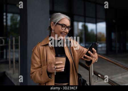 Eine Geschäftsfrau im Alter von 50 Jahren mit einer Tasse Kaffee schaut vor dem Hintergrund des Geschäftseingangs auf ein Mobiltelefon Stockfoto