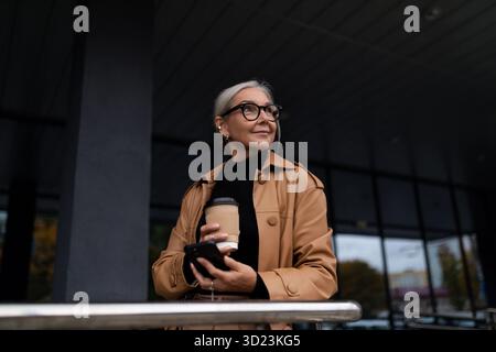 Reife grauhaarige kaukasische Geschäftsfrau mit einer Tasse Kaffee in der Nähe eines grauen Gebäudes draußen Stockfoto