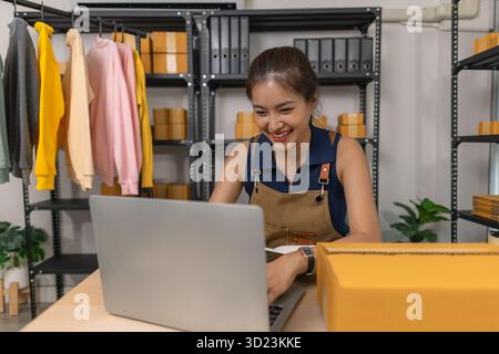 Junge Unternehmerin lächelt, während sie zu Hause an ihrem Laptop arbeitet, umgeben von Kleidung und Paketen. Sie leitet einen Online-Shop und kommuniziert mit wi Stockfoto
