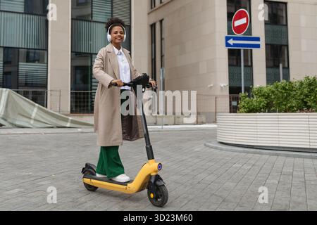 Stilvolle afroamerikanische Frau fährt einen Elektroroller vor dem Hintergrund eines Bürogebäudes Stockfoto
