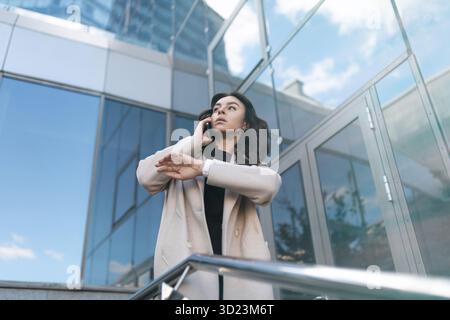 Erfolgreiche junge Geschäftsfrau, die auf einem Handy spricht und ihre Uhr im Hintergrund eines Bürogebäudes, Stron, ansieht Stockfoto