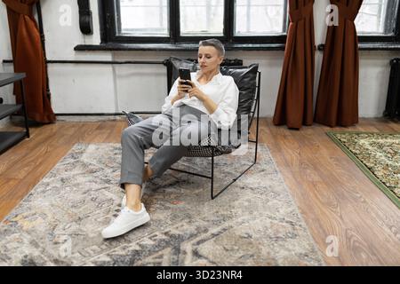 Weiße Frau mit kurzem Haarschnitt schaut auf den Bildschirm des Telefons, der auf einem Stuhl sitzt Stockfoto