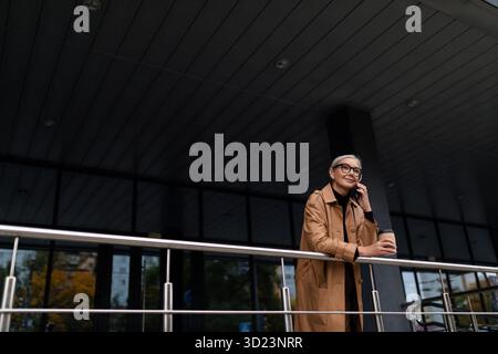 Stilvolle ältere Frau, die auf dem Handy in der Nähe des Bürogebäudes spricht Stockfoto