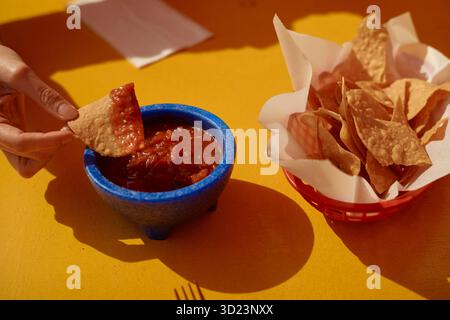 Tauch von Hand in Salsa, neben einem Korb mit Chips auf gelbem Tisch. Kalifornien, USA Stockfoto