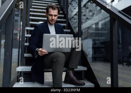 Geschäftsmann mittleren Alters in Geschäftskleidung arbeitet online auf einem Laptop, während er auf der Treppe neben dem Büro ce sitzt Stockfoto