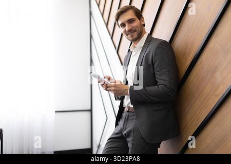 Ein erfolgreicher weißer Geschäftsmann mit einem Telefon in der Hand steht im Büro an der Wand Stockfoto