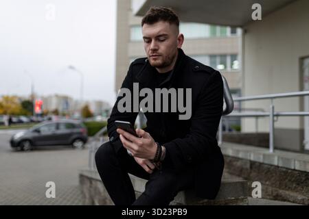 Brutaler junger Mann in einem schwarzen Mantel mit Handy auf dem Hintergrund des Bürozentrums Stockfoto