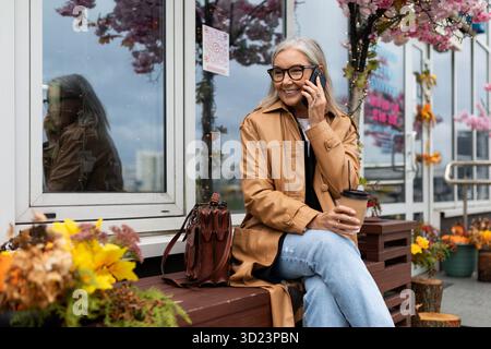 Stilvolle ältere Frau, die auf einem Mobiltelefon spricht und auf einer Bank neben einem Blumenladen sitzt Stockfoto