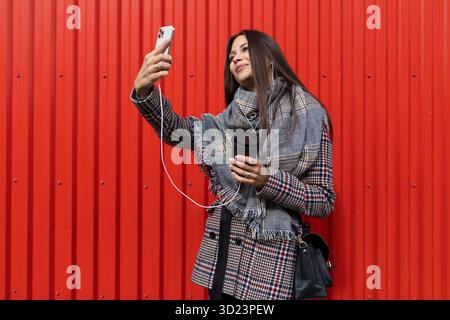Stilvolle junge Frau, die ein Selfie mit Kopfhörern in den Ohren und einer Tasse Kaffee in der Hand macht Stockfoto
