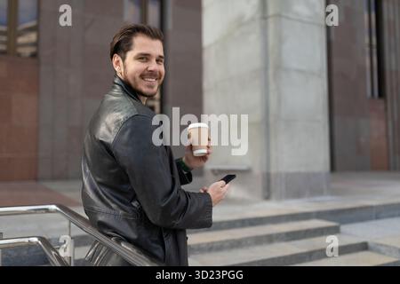 Erwachsener Mann mit einer Tasse Kaffee in den Händen und einem Handy draußen Stockfoto