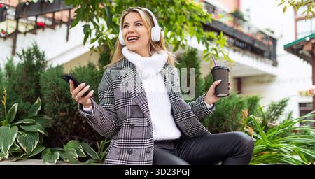 Erwachsene glückliche Frau während der Mittagspause in Kopfhörern, Musik hören und Kaffee trinken im Park vor dem Büro Stockfoto