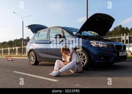 Eine junge Frau, die auf Hilfe wartet, sitzt in der Nähe eines kaputten Autos auf der Straße Stockfoto