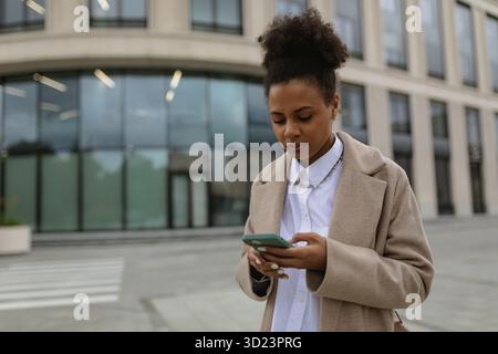 Junge afroamerikanische Geschäftsfrau, die auf einem Handy vor dem Hintergrund eines Business Centers in klassischer Kleidung schreibt Stockfoto