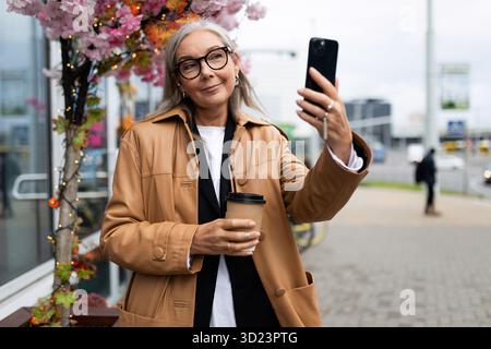 Stilvolle ältere Frau kommuniziert per Handy-Videoanruf in der Stadt mit einer Tasse Kaffee in der Hand Stockfoto