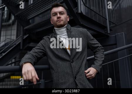 Stilvolles Porträt eines brutalen Mannes in einem Mantel auf dem Hintergrund eines schwarzen Gebäudes Stockfoto