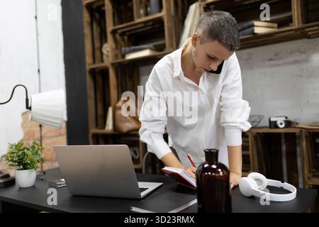 Eine Frau mit einem kurzen Haarschnitt arbeitet im Büro und spricht am Telefon und macht Notizen in einem Notizbuch Stockfoto
