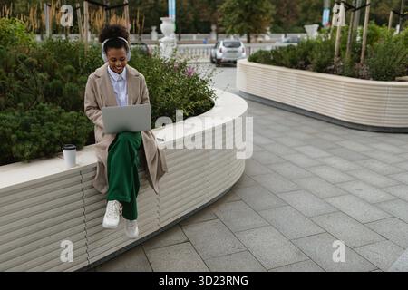 afroamerikanische Studentin, die außerhalb der Universität an einem Laptop arbeitet und Kopfhörer trägt, um Musik zu hören Stockfoto