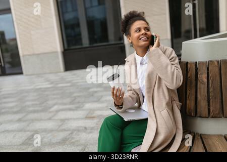 Porträt einer jungen afroamerikanischen Studentin, die auf einer Bank mit einem Laptop sitzt und auf einem Handy gegen das B spricht Stockfoto