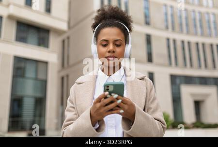 afroamerikanische stilvolle Frau in Kopfhörern auf dem Hintergrund eines Business Centers hält ein Telefon in ihren Händen Stockfoto