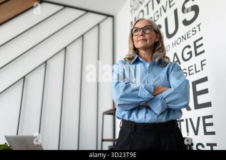 Die reife Geschäftsfrau steht im Büro mit überkreuzten Armen auf der Brust in einem blauen Hemd Stockfoto