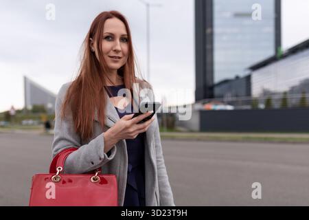 Rothaarige kaukasische Geschäftsfrau mit einem Telefon in der Hand auf dem Parkplatz des Bürozentrums Stockfoto
