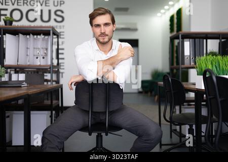 Junger Geschäftsmann mit ernsthaftem Ausdruck, der auf die Kamera blickt, während er hinten an vorne auf einem Stuhl im Büro sitzt Stockfoto
