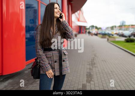 Schöne, stilvolle junge Frau, die auf einem Mobiltelefon mit einem Lächeln auf dem Gesicht vor dem Hintergrund eines rot-blauen Stadtgebäudes spricht Stockfoto