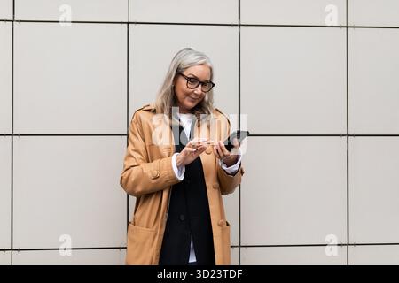 Stilvolle Geschäftsfrau 50 Jahre alt mit einem Mobiltelefon in den Händen vor dem Hintergrund einer beigefarbenen Wand draußen Stockfoto