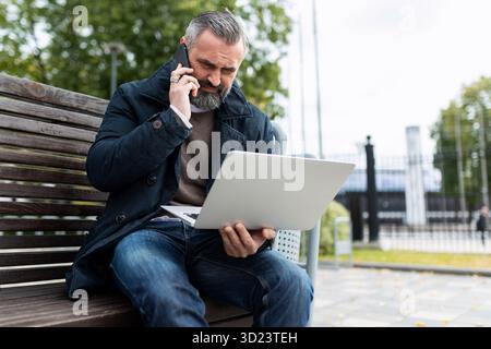 Auf dem Weg ins Büro spricht ein Mann am Telefon und schaut auf einen Laptop, während er auf einer Bank neben dem Business ce sitzt Stockfoto