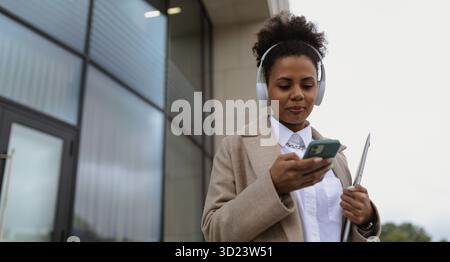afroamerikanische junge Frau in Kopfhörern mit Telefon und Laptop neben dem Bürogebäude Stockfoto