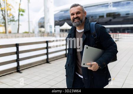 Reifer Mann mit Laptop und Rucksack, der sich lächelnd in die Kamera schaut Stockfoto