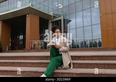 afroamerikanische Studentin mit Laptop sitzt auf den Stufen vor dem Verwaltungsgebäude Stockfoto
