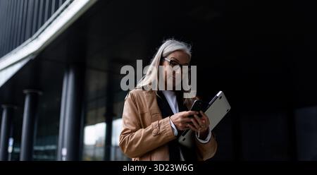 Eine reife Geschäftsfrau geht mit einem Laptop und einem Telefon in der Hand ins Büro Stockfoto