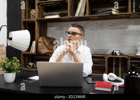 Moderne Geschäftsfrau in Brille mit kurzer Frisur sitzt an ihrem Schreibtisch und denkt an einen Laptop Stockfoto