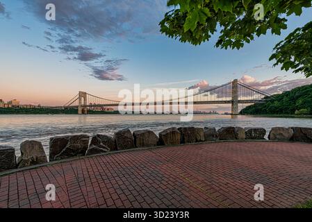 Majestätische Hängebrücke über einen breiten Fluss bei Sonnenuntergang mit einer Skyline der Stadt in der Ferne. New York City, NY, USA George Washington Bridge, Hudson River Stockfoto