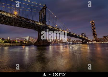 Beleuchtete Stadtlandschaft in der Abenddämmerung mit Brooklyn Bridge, die sich über einen ruhigen Fluss erstreckt. New York City, NY, USA Stockfoto