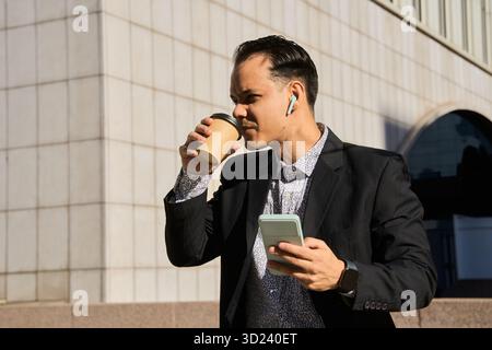 Junger lateinamerikanischer Geschäftsmann in Anzug trinkt Kaffee und überprüft das Smartphone draußen, Multitasking mit schnurlosen Ohrhörern in urbaner Umgebung Stockfoto