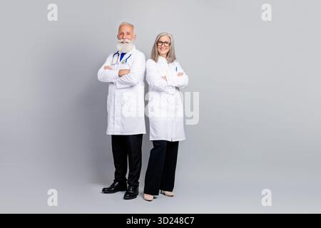 Zwei erfahrene Ärzte stehen zusammen in weißen Mänteln und lächeln selbstbewusst für ein professionelles medizinisches Porträt vor grauem Hintergrund Stockfoto