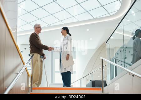 Älterer kaukasischer Mann mit Stock schüttelnd die Hände mit jungen Erwachsenen asiatischen Frau Doktor, der in moderner medizinischer Einrichtung steht, sowohl lächelnd als auch Augenkontakt auf Treppe, nach medizinischem Termin Stockfoto