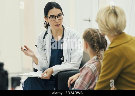 Kaukasische Ärztin, die während der Konsultation im klinischen Setting zusammen sitzende kaukasische Frau mittleren Alters und kaukasisches Kind medizinische Informationen erklärt Stockfoto