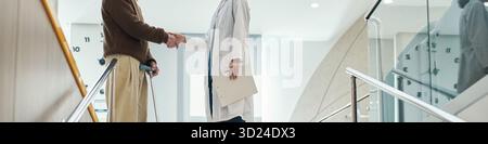 Kopfzeile eines Mannes mittleren Alters mit Behinderung mit Krücken, die Hand schütteln mit einem jungen erwachsenen weiblichen Arzt im weißen Labormantel, stehend in einer modernen medizinischen Einrichtung in der Nähe einer Treppe Stockfoto