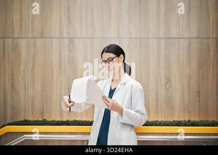 Kaukasische junge Ärztin mit Brille, die Dokumente überprüft, während sie in moderner Büroumgebung steht, Papiere hält und Informationen mit fokussiertem Ausdruck untersucht Stockfoto