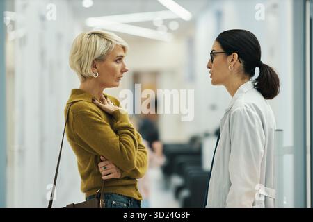 Kaukasische Frau mittleren Alters spricht mit jungen Erwachsenen asiatischen Ärztin in der medizinischen Einrichtung Flur, beide stehend und gegenüberstehend, diskutieren gesundheitliche Bedenken Stockfoto