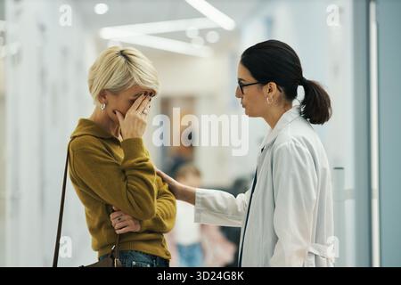 Kaukasische Frau mittleren Alters, die das Gesicht mit der Hand bedeckt, während sie im Flur steht und von einer jungen, erwachsenen kaukasischen Ärztin getröstet wird, die Brille und Laborkittel trägt Stockfoto