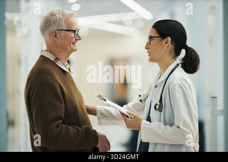 Älterer kaukasischer Mann lächelt, während er mit einem jungen Erwachsenen kaukasischen weiblichen Doktor spricht, der ein Klemmbrett hält, zusammen in einer modernen medizinischen Einrichtung steht und sich in einem freundlichen Gespräch befindet Stockfoto