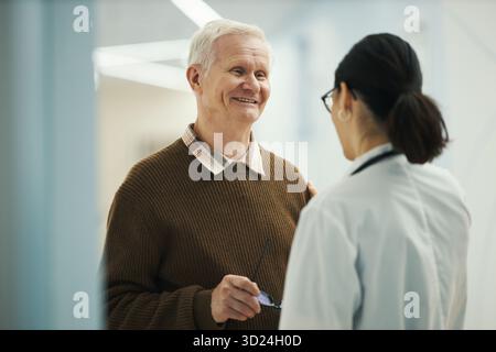 Älterer kaukasischer Mann lächelt, während er mit jungen erwachsenen Frau Arzt in der medizinischen Einrichtung spricht, Brille in der Hand hält, in freundlichem Gespräch während der Konsultation Stockfoto